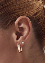 A close-up of a person's ear wearing a gold huggie hoop earring and a small round bezel stud earring made for solid 14k gold with white topaz fro SB Studio