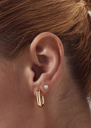 A close-up of a person's ear wearing a gold huggie hoop earring and a small round bezel stud earring made for solid 14k gold with white topaz fro SB Studio