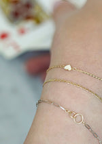 SB Studio 14k yellow gold heart charm bracelet with white topaz stones worn on a wrist with other gold chains.